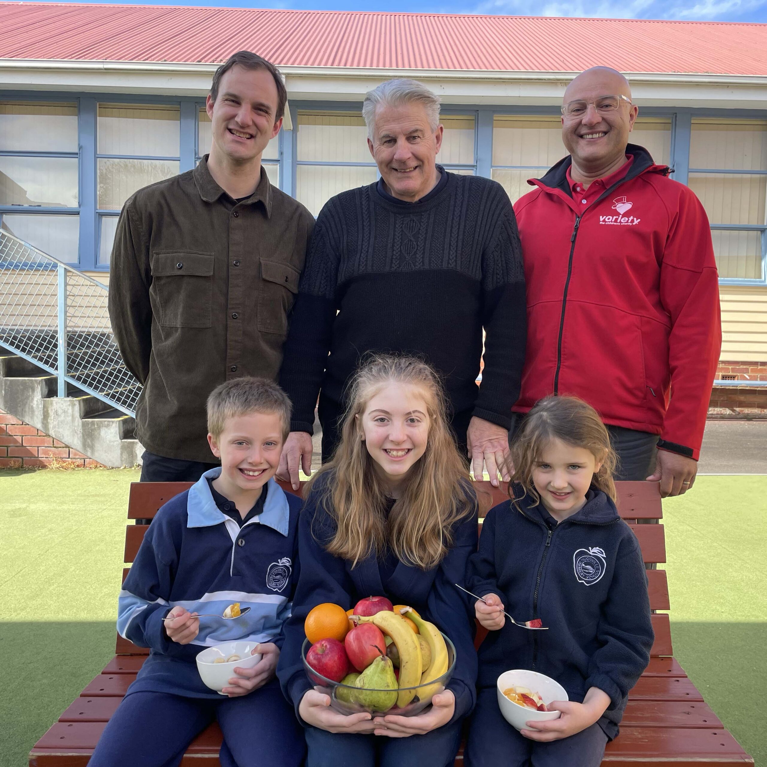 3 adult men who are the principal, teacher and Variety representative standing behind two young girls with brown hair and one boy about 10 years old who are holding a bowl of fruit and cereal