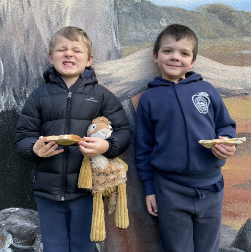 two young boys in school uniform in front of a mural eating toast and smiling