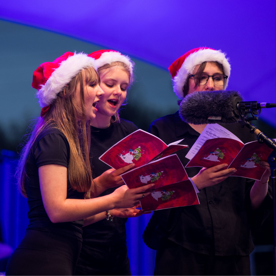 three girls in santa hats singing Christmas Carols into a microphone