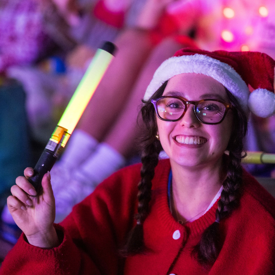 young woman in glasses and a santa hat with a big smile holding a light up glow stick